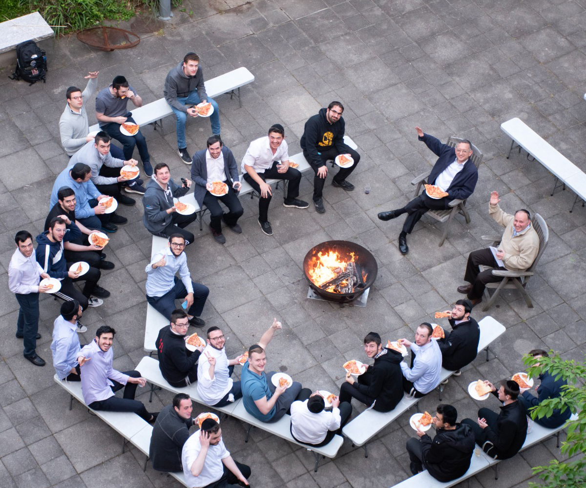 Overhead view of Professor Chayim Herskowitz and Professor Newman sitting around and outdoor fire pit with various LCM students