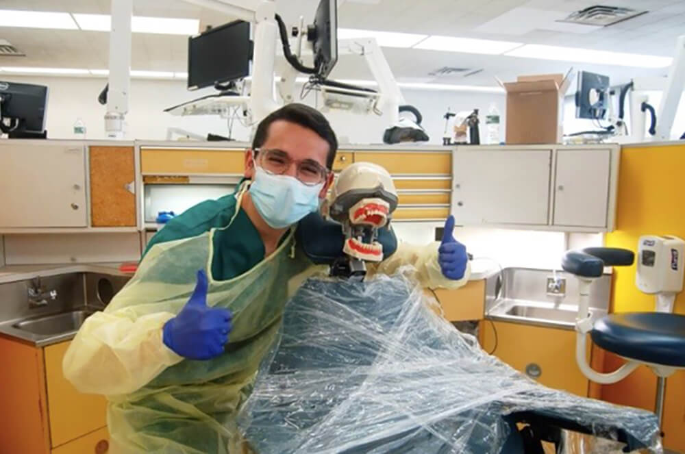 Mendy Antelis in scrubs, mask, and globes, giving a thumbs up to the camera with his arm around a dental chair, at rutgers dental school of medicine. In the dental chair is a dental school training manikin of head and teeth.