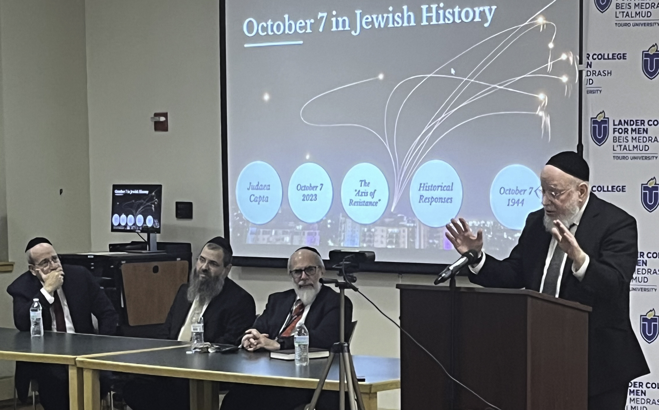 Rabbi Sacks at a podium on the right and the rest of Touro/LCM leadership at a table at the front of the room in front of a screen that says "October 7 in Jewish History"