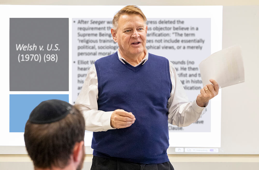 Professor Tom Rozinski standing in front of classroom in front of whiteboard in white shirt and blue vest.
