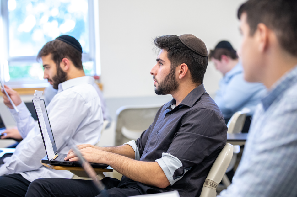 Male students in a classroom seated at a desk with a laptop wearing yarmulkes.
